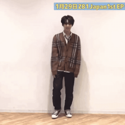 A Man Wearing A Plaid Cardigan And Black Jeans Stands In Front Of A Wall That Says Japan 1St Ep GIF