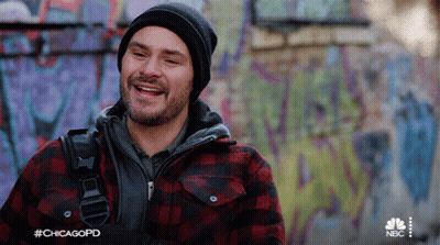 A Man Wearing A Plaid Jacket And A Beanie Is Smiling In Front Of A Graffiti Covered Wall .. GIF