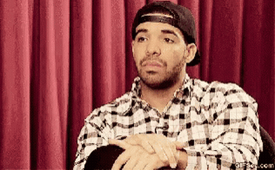 A Man Wearing A Plaid Shirt And A Baseball Cap Is Sitting In Front Of A Red Curtain GIF