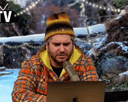 A Man Wearing A Plaid Shirt And A Beanie Is Sitting In Front Of An Apple Laptop GIF