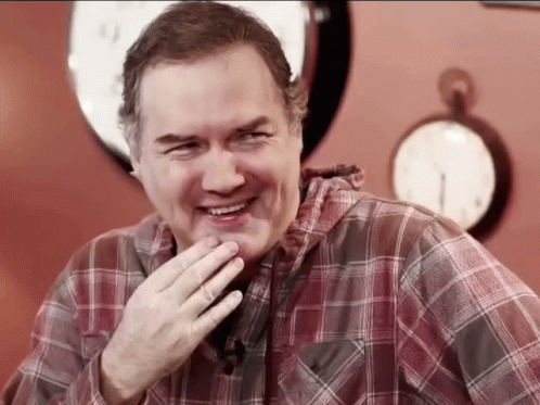 A Man Wearing A Plaid Shirt Is Laughing In Front Of A Clock GIF