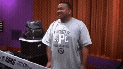 A Man Wearing A Property Of Pl Shirt Stands In Front Of A Korg Triton Keyboard GIF