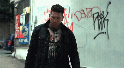 A Man Wearing A Punisher T-Shirt Stands In Front Of A Graffiti Covered Wall GIF