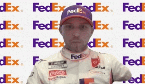 A Man Wearing A Purple Fedex Hat Stands In Front Of A Fedex Logo GIF