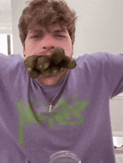 A Man Wearing A Purple Shirt With A Green Letter K On It Is Eating Pickles GIF