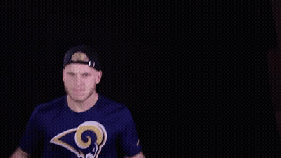 A Man Wearing A Rams T-Shirt And A Hat Is Holding His Hands Up In The Air . GIF