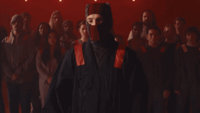 A Man Wearing A Red And Black Mask Stands In Front Of A Crowd Of People GIF