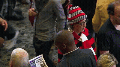 A Man Wearing A Red And Black Striped Hat Is Reading A Newspaper In A Crowd Of People GIF