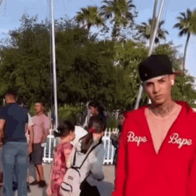 A Man Wearing A Red Bape Hoodie And A Black Hat Is Standing In Front Of A Crowd Of People . GIF