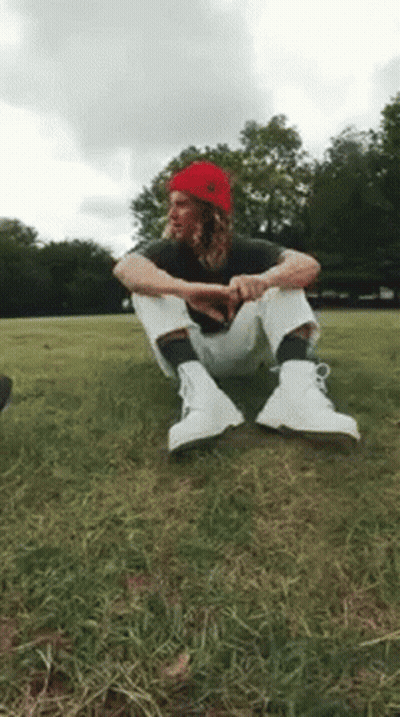 A Man Wearing A Red Beanie And White Boots Is Sitting On The Grass . GIF