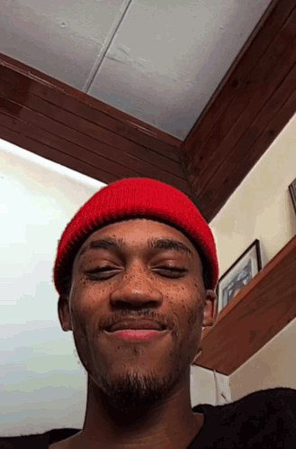 A Man Wearing A Red Beanie Looks At The Camera With His Eyes Closed GIF