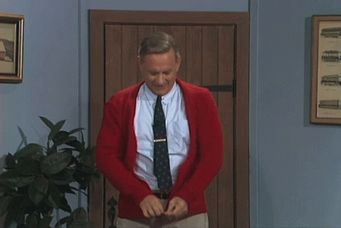 A Man Wearing A Red Cardigan And Tie Is Standing In Front Of A Wooden Door GIF