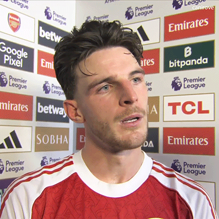A Man Wearing A Red Emirates Jersey Stands In Front Of A Wall Of Advertisements GIF