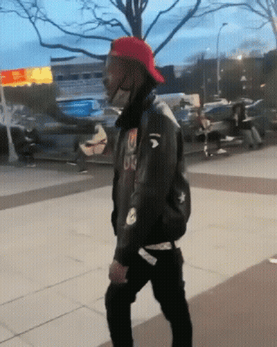 A Man Wearing A Red Hat And A Black Jacket With The Letter U On It Is Walking Down A Sidewalk GIF