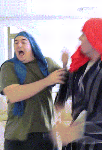 A Man Wearing A Red Hat And A Blue Scarf Stands Next To Another Man GIF