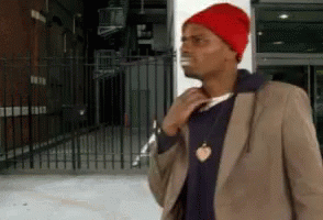 A Man Wearing A Red Hat And A Brown Jacket Is Standing In Front Of A Building GIF
