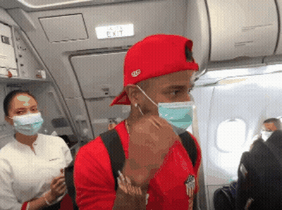 A Man Wearing A Red Hat And A Face Mask Stands In Front Of An Airplane Exit Sign GIF