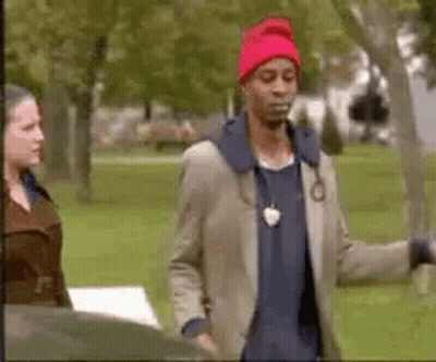 A Man Wearing A Red Hat And A Jacket Is Standing In A Park With A Woman . GIF