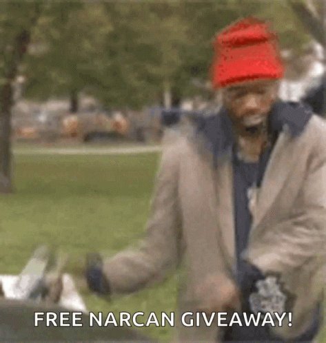 A Man Wearing A Red Hat And A Jacket Is Standing In A Park With The Words `` Free Narcan Giveaway ! '' GIF