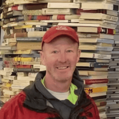 A Man Wearing A Red Hat And A Jacket Stands In Front Of A Pile Of Books GIF