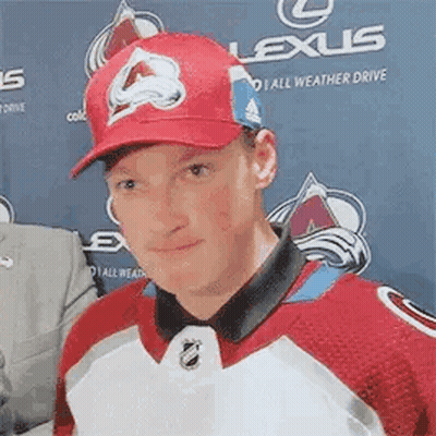 A Man Wearing A Red Hat And A Red And White Hockey Jersey Is Standing In Front Of A Lexus Banner . GIF