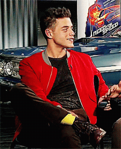 A Man Wearing A Red Jacket Sits In Front Of A Need For Speed Poster GIF