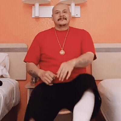 A Man Wearing A Red Shirt And A Gold Necklace Is Sitting On A Chair GIF
