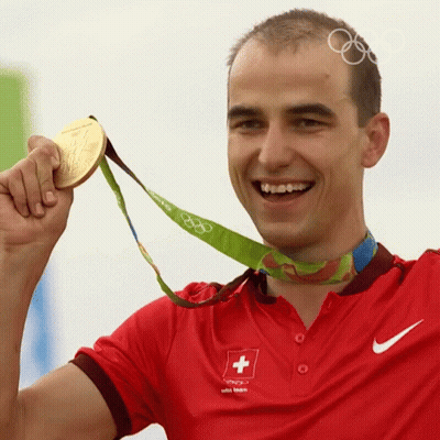 A Man Wearing A Red Shirt With A Swiss Cross On It Holds Up A Gold Medal GIF