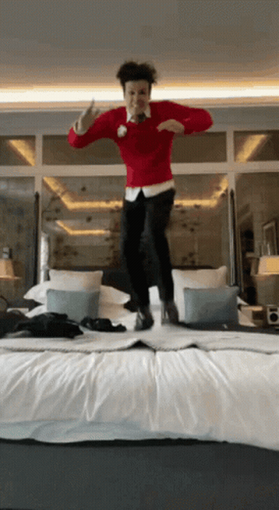 A Man Wearing A Red Sweater Is Jumping On A Bed GIF
