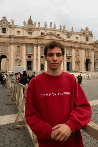 A Man Wearing A Red Tommy Hilfiger Shirt Stands In Front Of A Large Building GIF