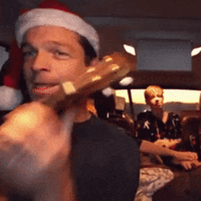 A Man Wearing A Santa Hat Is Holding A Guitar In His Hand GIF