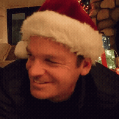 A Man Wearing A Santa Hat Is Smiling In A Living Room . GIF