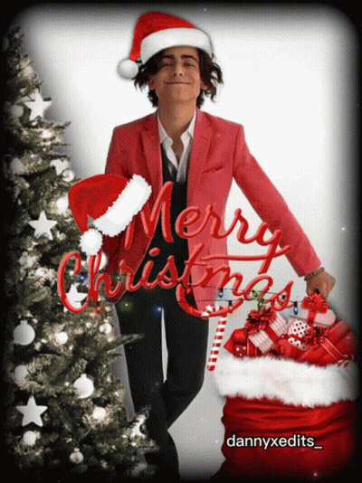 A Man Wearing A Santa Hat Is Standing Next To A Christmas Tree With Merry Christmas Written In Red Letters GIF
