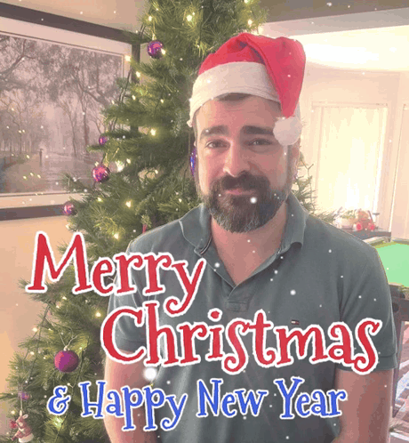 A Man Wearing A Santa Hat Stands In Front Of A Christmas Tree GIF