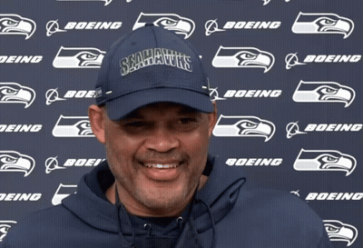 A Man Wearing A Seahawks Hat Stands In Front Of A Boeing Logo GIF