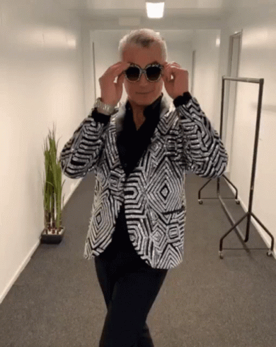 A Man Wearing A Sequined Jacket And Sunglasses Stands In A Hallway GIF