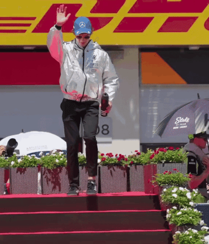 A Man Wearing A Silver Jacket And A Blue Hat Stands On A Stage In Front Of Dhl GIF