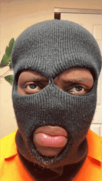 A Man Wearing A Ski Mask And An Orange Shirt Looks At The Camera GIF