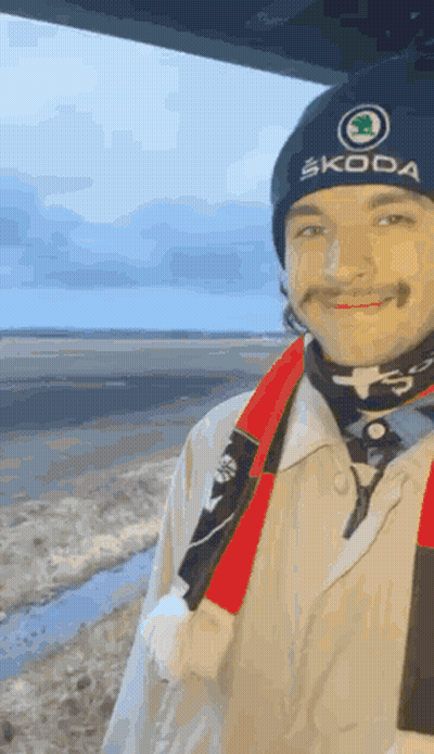 A Man Wearing A Skoda Hat And Scarf Is Standing In Front Of A Body Of Water . GIF