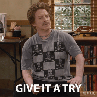 A Man Wearing A Skull And Crossbones Shirt Is Sitting In Front Of A Bookshelf And Says Give It A Try GIF