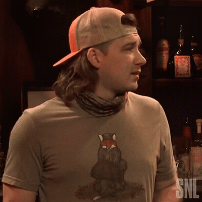 A Man Wearing A Snl Shirt Stands In Front Of A Shelf Full Of Liquor Bottles GIF