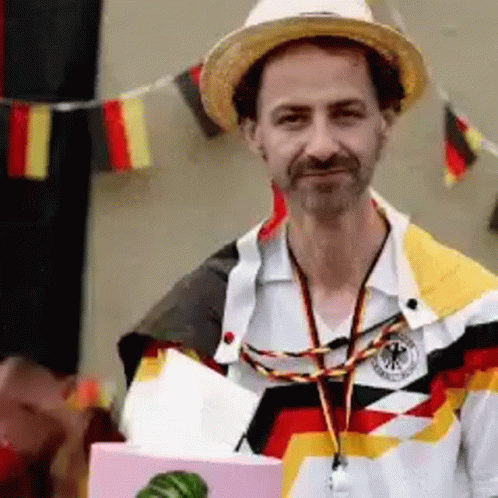 A Man Wearing A Straw Hat And A German Shirt Is Holding A Box GIF