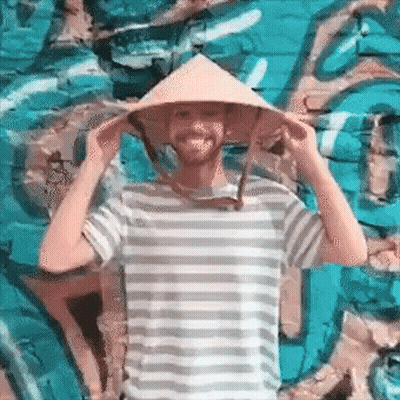 A Man Wearing A Striped Shirt And A Conical Hat Is Standing In Front Of A Blue Wall . GIF