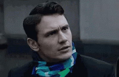 A Man Wearing A Suit And A Scarf Around His Neck Is Looking At The Camera . GIF