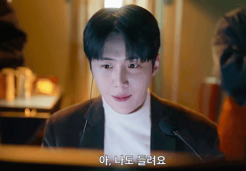 A Man Wearing A Suit And A White Turtleneck Talks Into A Microphone With Korean Writing On The Bottom Right GIF