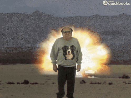 A Man Wearing A Sweater With A Dog On It Is Standing In Front Of An Explosion Sponsored By Quickbooks GIF