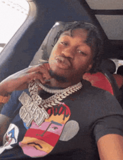 A Man Wearing A T-Shirt With A Face On It Is Sitting In A Car With His Hand On His Chin GIF