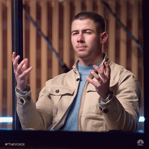 A Man Wearing A Tan Jacket And A Blue Sweater Is Holding His Hands Up In The Air GIF
