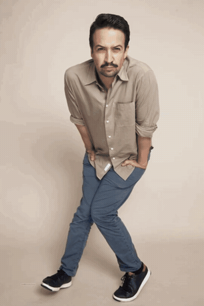 A Man Wearing A Tan Shirt And Blue Pants Is Standing With His Hands In His Pockets GIF
