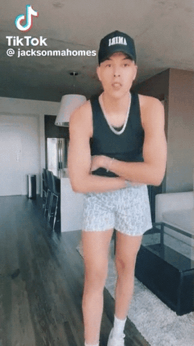 A Man Wearing A Tank Top And Shorts Is Standing In A Living Room With His Arms Crossed . GIF